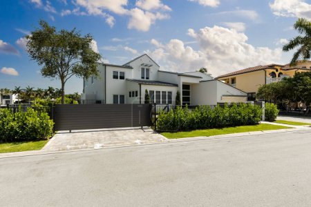 Téléchargez les photos : Vue de face d'une élégante maison blanche moderne avec passerelle menant à la porte d'entrée, garage, herbe courte, arbres, palmiers, bordure privée, trottoir, ciel bleu - en image libre de droit
