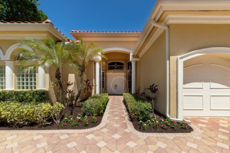 Facade of elegant colonial-style mansion in Boca Raton, with tropical front garden, cobblestone driveway, palms, trees, short grass, sidewalk, blue sky