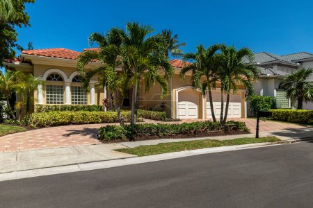 Facade of elegant colonial-style mansion in Boca Raton, with tropical front garden, cobblestone driveway, palms, trees, short grass, sidewalk, blue sky
