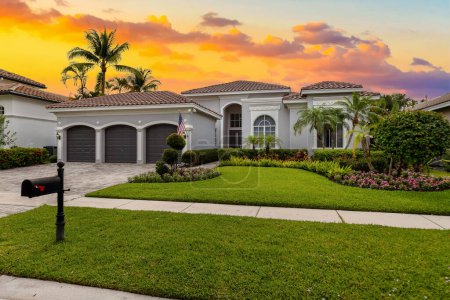 Fassade eines eleganten Hauses mit grauen Wänden mit weißen Details, einem roten Ziegeldach, einem Vorgarten mit üppigen tropischen Pflanzen, Palmen, kurzem Gras, Bürgersteig, Einfahrt und Garage