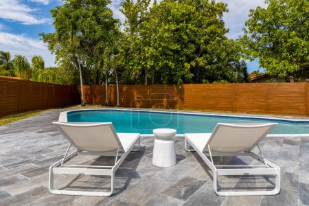 Téléchargez les photos : Vue sur la piscine turquoise avec deux chaises longues, clôture en bois, palmiers et arbres, ciel bleu - en image libre de droit