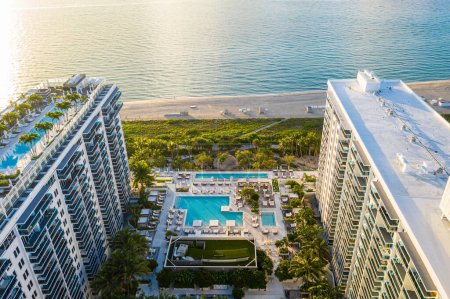 Aerial drone shot of Miami Beach coastline with towers and modern buildings, blue sky, bay, waves, beach, tropical vegetation, swimming pools