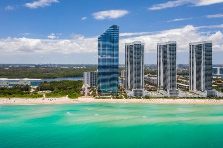 Aerial drone shot of Sunny Isles Beach city coastline with modern towers, beach, bay blue sky with clouds in the background, cityscape, swimming pools, turquoise sea