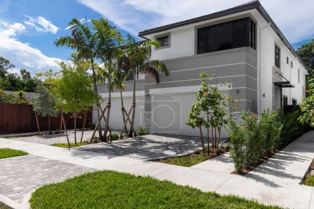 Téléchargez les photos : Facade of elegant and modern house in white and gray tones in the Tarpon River neighborhood in Fort Lauderdale, driveway, garage, sidewalk, short grass, palm trees, path to the front door, blue sky and tropical climate - en image libre de droit