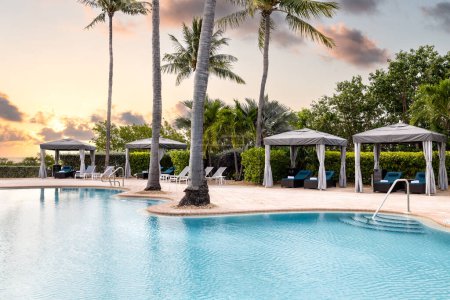 Téléchargez les photos : Beau coucher de soleil dans une piscine d'hôtel, avec chaises longues avec gazebos, ventilateurs, palmiers, ciel orange et bleu, vue relaxante - en image libre de droit