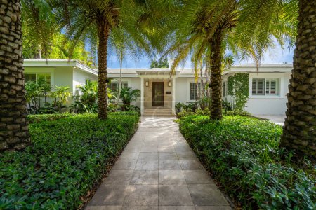 facade of a modern and luxurious colonial-style house in the city of Coral Gables, Miami-Dade, USA, with large tropical vegetation around it, open garage, main entrance, sidewalk, short grass, privet flower beds
