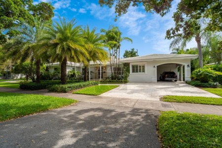 facade of a modern and luxurious colonial-style house in the city of Coral Gables, Miami-Dade, USA, with large tropical vegetation around it, open garage, main entrance, sidewalk, short grass, privet flower beds