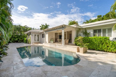 Beautiful backyard with concrete floor and pool, surrounded by tropical plants, porch with sun loungers, outdoor furniture, vines, located in Coral Gables