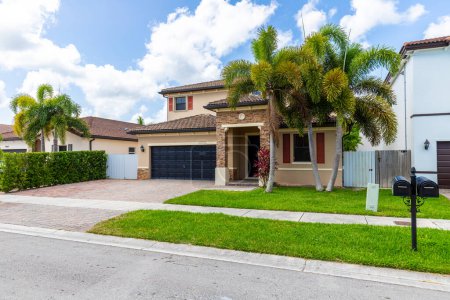 Photo for Front of house in beige and navy colors, with stone details, red tiled roof, located in Miami-Dade, FL, USA, classic suburban style houses, blue sky, outdoor furniture, front view the lagoon, trees. - Royalty Free Image