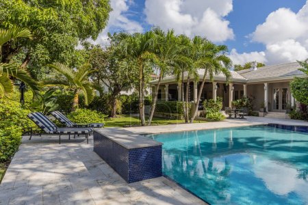 Backyard of elegant and luxurious mansion, with palms and lots of plants around, beautiful pool, covered patio, outdoor furniture, shortgrass, basketball court and blue sky with clouds