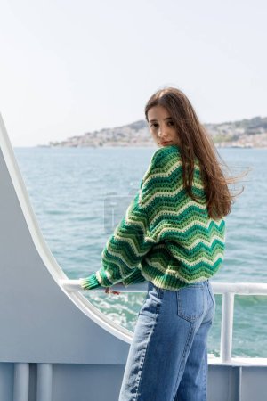 Young woman in knitted sweater and jeans looking at camera while standing on ferry boat with Princess islands at background