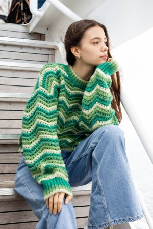 Young woman in sweater and jeans looking away while sitting on stairs on yacht 