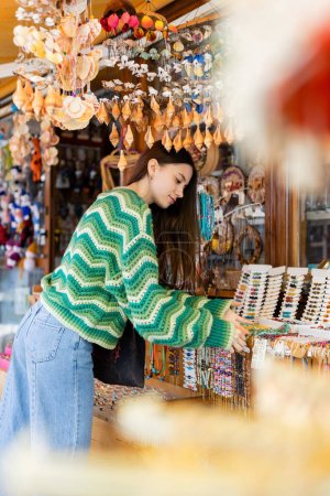 Photo for Side view of brunette woman in knitted sweater choosing accessories on market in Turkey - Royalty Free Image