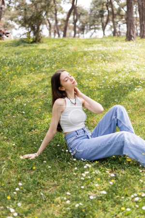 Young woman in top and jeans touching neck while sitting on lawn with flowers in park 