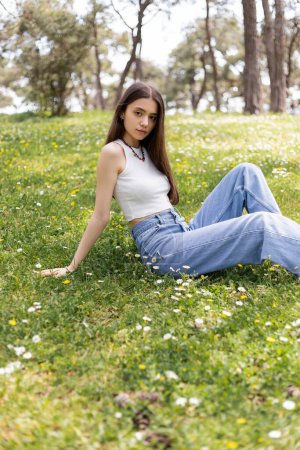 Young brunette woman in top and jeans looking at camera while sitting on lawn with flowers in park 