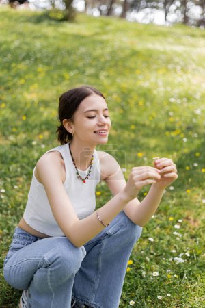 Smiling young woman in top and jeans holding daisy flower on lawn in park 