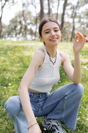Téléchargez les photos : Jeune femme souriante en haut et jeans regardant caméra sur prairie floue avec des fleurs dans le parc - en image libre de droit