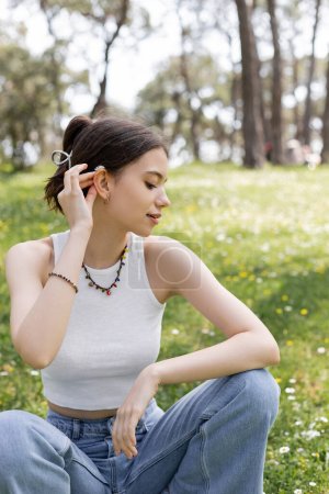 Téléchargez les photos : Vue latérale de la jeune femme mettant fleur de marguerite dans les cheveux tout en étant assis sur la pelouse dans le parc - en image libre de droit