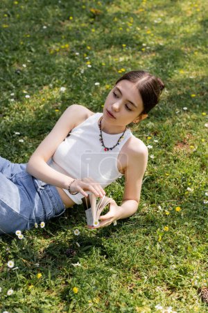 Hochwinkelaufnahme der jungen brünetten Frau in Top und Jeans, die Buch hält, während sie im Park auf dem Rasen mit Gänseblümchen liegt 