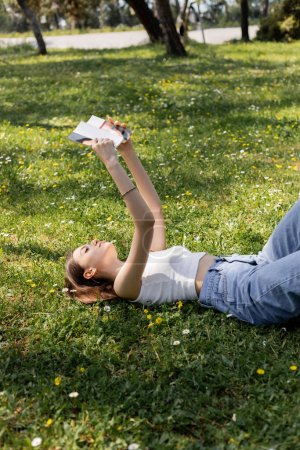 Joven morena en la parte superior y jeans libro de lectura mientras está acostado en el prado con flores en el parque 
