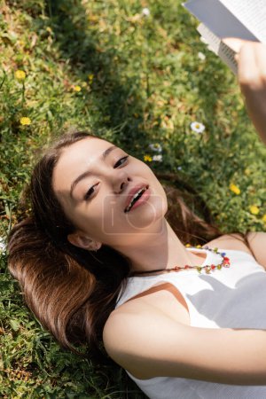 Blick aus der Vogelperspektive auf eine lächelnde junge Frau, die in die Kamera blickt, während sie Buch in der Hand hält und auf einer Blumenwiese im Park liegt 