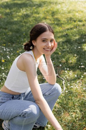 Téléchargez les photos : Souriant jeune femme avec fleur dans les cheveux regardant la caméra sur la pelouse dans le parc - en image libre de droit