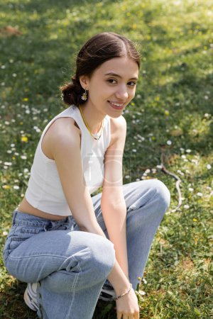 Photo for Carefree young woman in top and jeans looking at camera on meadow with flowers in park - Royalty Free Image