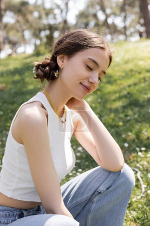 Téléchargez les photos : Joyeux jeune femme avec des fleurs dans les cheveux assis sur la pelouse floue dans le parc d'été - en image libre de droit
