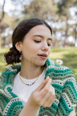 Young brunette woman with knitted sweater on shoulders holding blurred daisy flower in park 