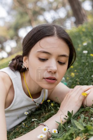 Young woman in top looking at flowers while lying on meadow in park 