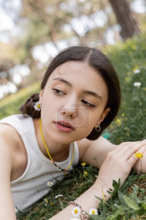 Brunette woman in top looking away while lying on lawn with flowers in summer park 