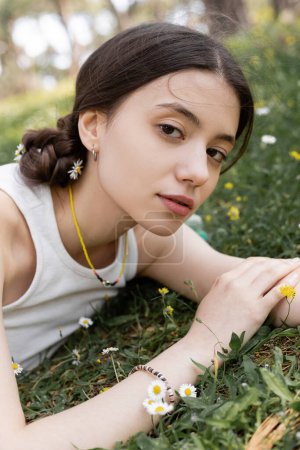 Porträt einer jungen brünetten Frau im Top, die in die Kamera schaut, in der Nähe von Gänseblümchen auf einer Wiese im Park 