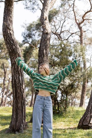 Foto de Mujer joven en suéter de punto y jeans mirando hacia otro lado mientras está de pie en el parque de verano - Imagen libre de derechos