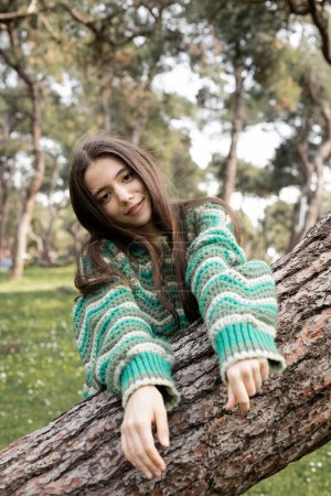 Portrait of smiling young woman in knitted sweater looking at camera near tree in summer park 