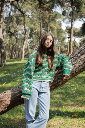 Brunette woman in knitted sweater and jeans looking at camera near tree in park 