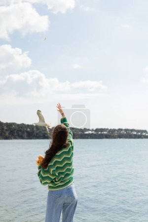 Back view of young woman holding bread near seagull flying over sea in Istanbul