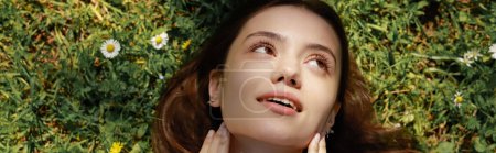Photo for Top view of positive brunette woman touching face while lying on meadow with flowers, banner - Royalty Free Image