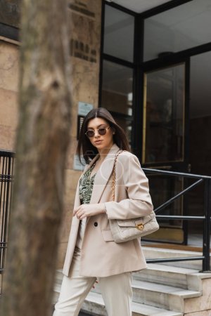 young brunette woman in stylish sunglasses and trendy outfit with white pants and beige blazer walking with handbag near modern building and blurred tree trunk on street in Istanbul 