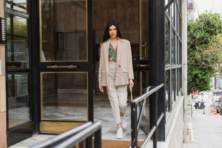 young brunette woman in trendy outfit with white pants, blouse and beige blazer walking out of modern building while holding handbag with chain strap on street in Istanbul 