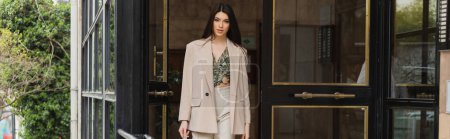 Photo for Young brunette woman in trendy outfit with white pants, blouse and beige blazer walking out of modern building on urban street in Istanbul, banner, entrance door - Royalty Free Image