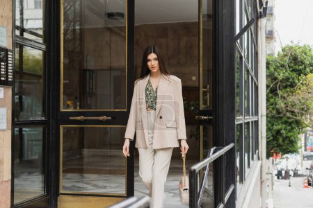 Photo for Brunette and young woman in trendy outfit with white pants, blouse and beige blazer walking out of modern building while holding handbag with chain strap on street in Istanbul - Royalty Free Image