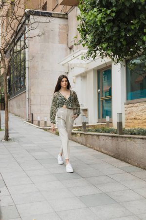 mujer joven con pelo largo en traje de moda con pantalones beige, blusa recortada y bolso con correa de cadena caminando con la mano en el bolsillo cerca de edificio moderno y árbol verde en la calle en Estambul 
