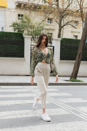 stylish woman with brunette long hair in trendy outfit with beige pants, cropped blouse and handbag with chain strap walking on crosswalk of urban street in Istanbul, blurred house on background 