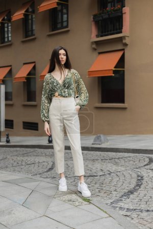 Photo for Stylish woman with brunette long hair in trendy outfit with beige pants, cropped blouse and handbag with chain strap standing with hand in pocket near blurred building on urban street in Istanbul - Royalty Free Image