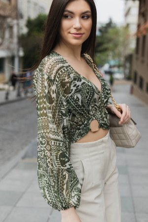 mujer elegante con pelo largo morena en traje de moda con pantalones beige, blusa recortada y bolso con correa de cadena de pie y sonriendo en la calle urbana de Estambul 