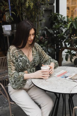 mujer joven con el pelo largo morena y maquillaje sentado en la silla cerca de mesa redonda bistro con periódico y la celebración de la taza de papel con café cerca de plantas borrosas en la terraza de la cafetería en Estambul 