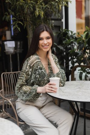 joyous young woman with long brunette hair and makeup sitting on chair near round bistro table and holding coffee in paper cup and smiling near blurred plants on terrace of cafe in Istanbul