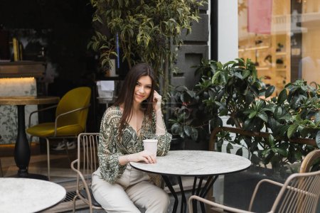 smiling woman with long brunette hair and makeup sitting on chair near round bistro table and holding coffee in paper cup near blurred plants on terrace of cafe in Istanbul 