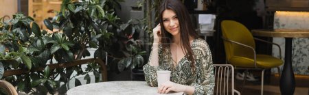 sonriente joven con el pelo largo morena sentado en la silla cerca de mesa redonda bistro y la celebración de café en taza de papel cerca de plantas borrosas en la terraza de la cafetería en Estambul, pancarta 