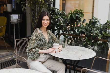 Charmante junge Frau mit langen brünetten Haaren sitzt auf einem Stuhl in der Nähe des runden Bistrotisches und hält Kaffee in einer Papptasse, während sie wegschaut und neben verschwommenen Pflanzen auf der Terrasse des Cafés in Istanbul lächelt 